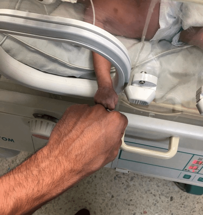 A premature baby in a NICU incubator reaches an arm through an opening to touch an adult's fist.