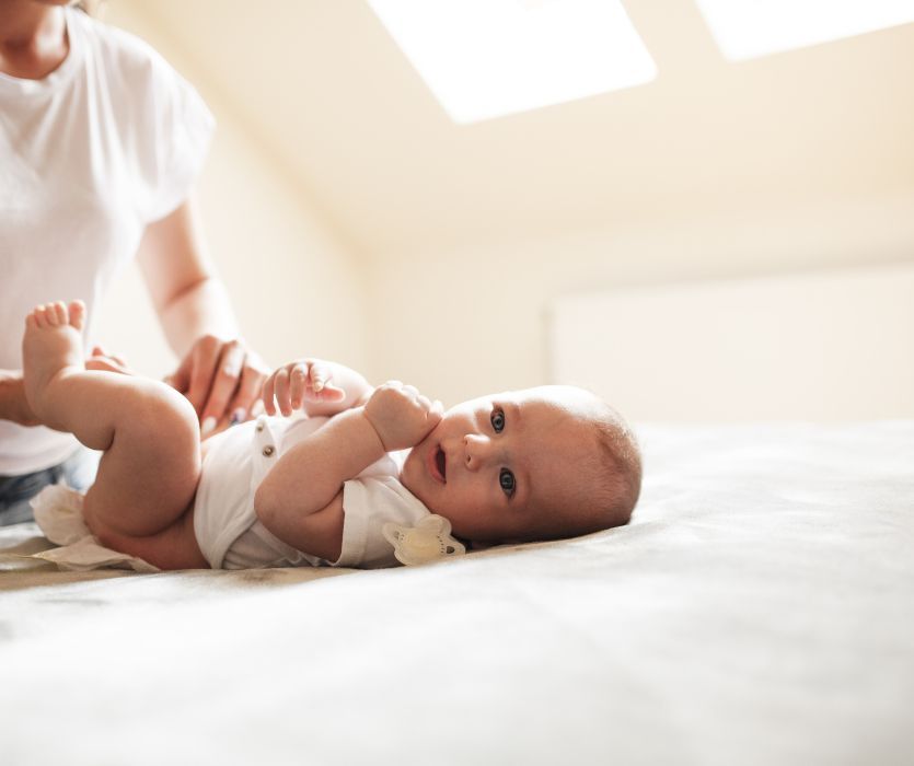 A baby in a white bodysuit lying on a bed while a caregiver changes their diaper.