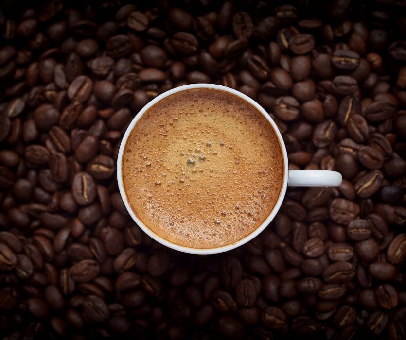 Top-down view of a white cup of coffee with crema foam resting on a bed of roasted coffee beans.