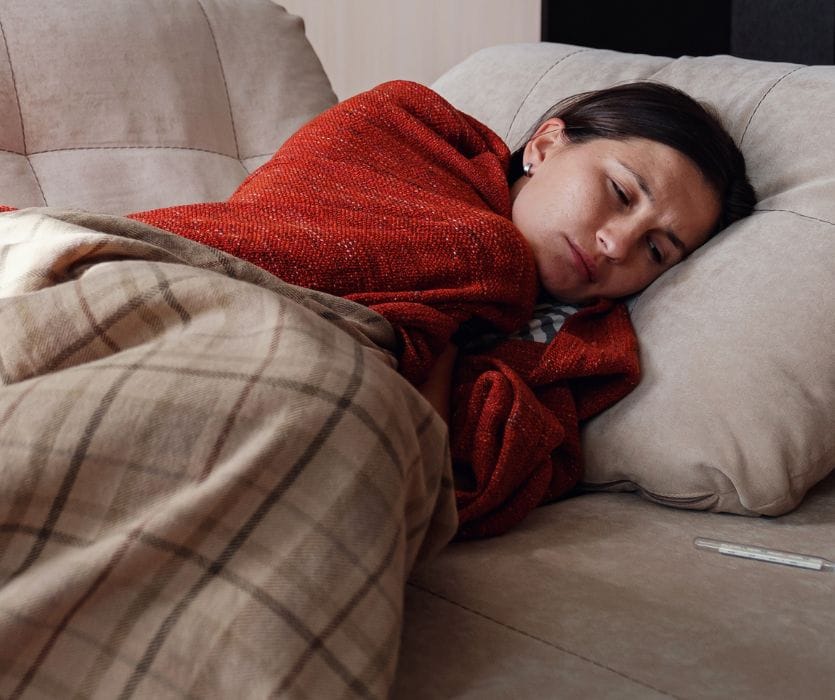 A woman looks unwell while lying on a sofa wrapped in blankets next to a thermometer.