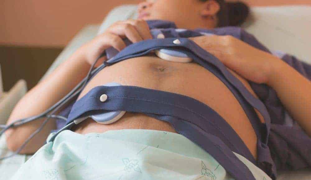 Fetal heart rate monitors strapped to the belly of a pregnant person lying in a hospital bed.