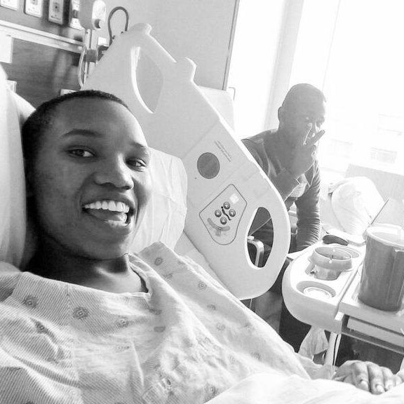 Black and white photo of a smiling person in a hospital bed while a man smiles and makes a peace sign in the background.