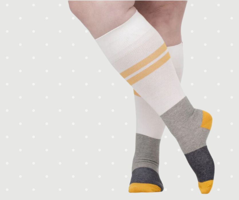 White knee-high compression socks with yellow stripes, grey heels, and dark grey toes on a person’s crossed legs.