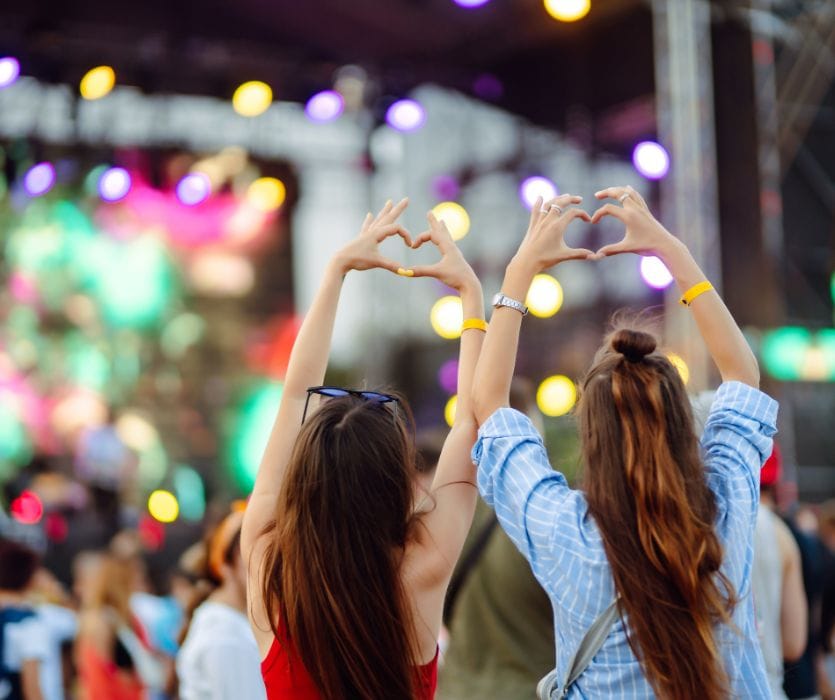 Two women with their backs to the camera make heart shapes with their hands at an outdoor concert with bright stage lights.