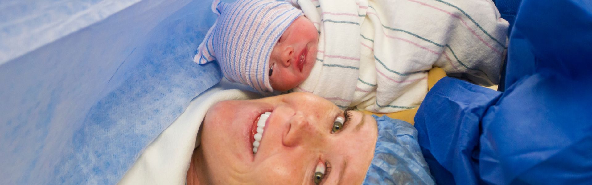 Smiling woman in surgical cap laying next to her newborn baby in a hospital swaddle.