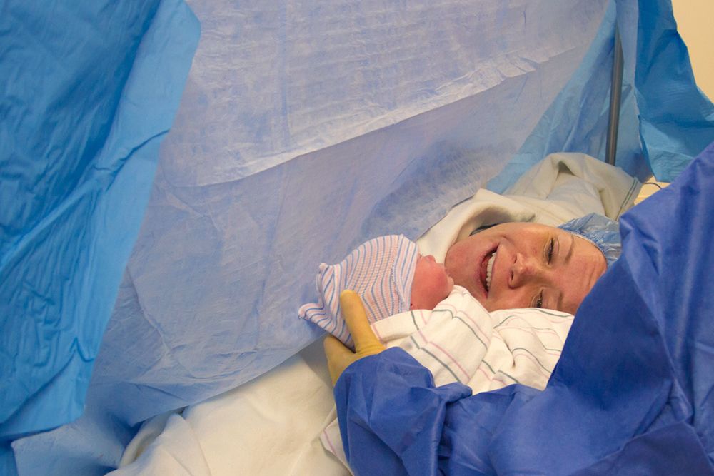 Smiling mother behind a surgical drape meeting her newborn baby for the first time during a C-section.
