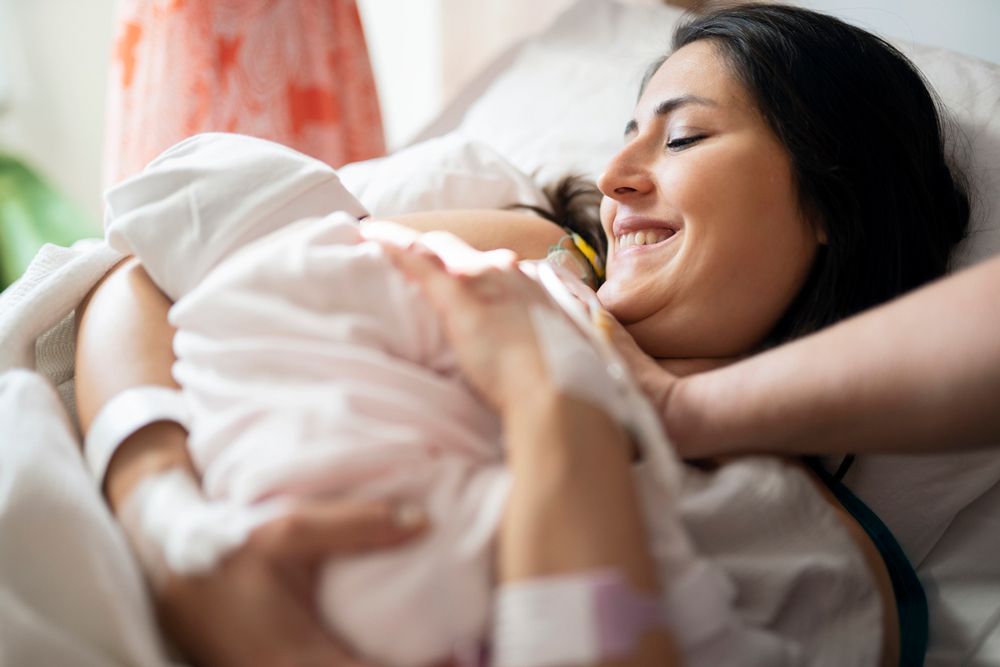 A smiling mother lying in a hospital bed holds her newborn baby wrapped in a white blanket to her chest.