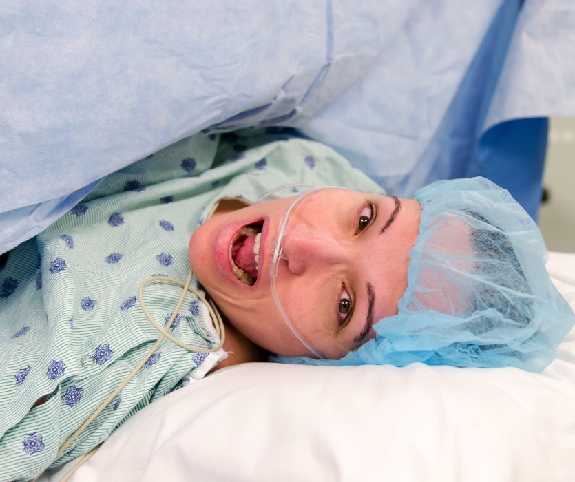 A patient in a surgical cap and gown with an oxygen nasal cannula lies on an operating table behind a blue curtain.