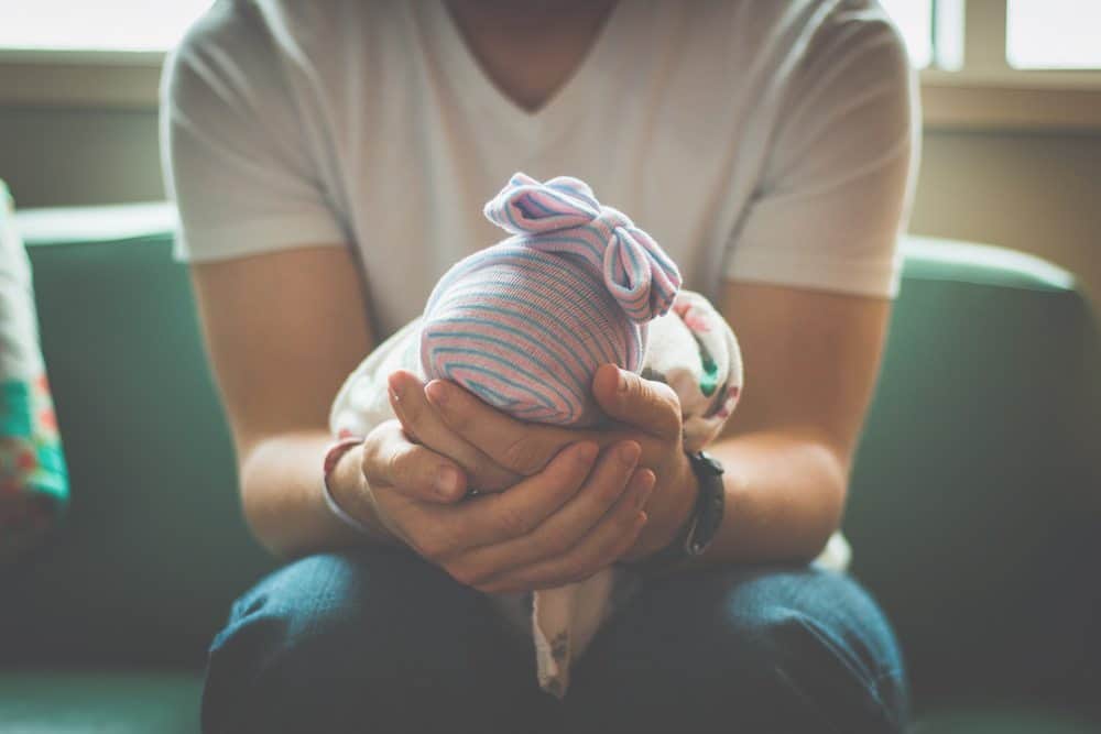 A person’s hands gently cradling the head of an infant wearing a blue and pink striped hat with a bow.