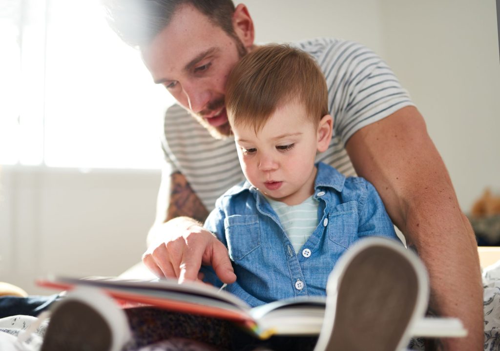 A father points at a book while reading with his young son indoors.