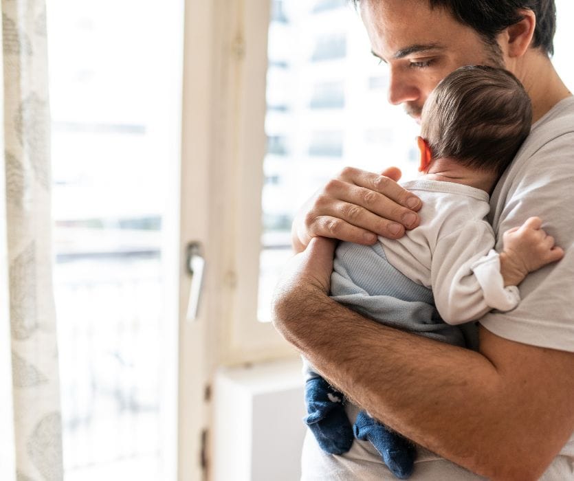 A man with a somber expression holds an infant close to his chest while standing by a window.