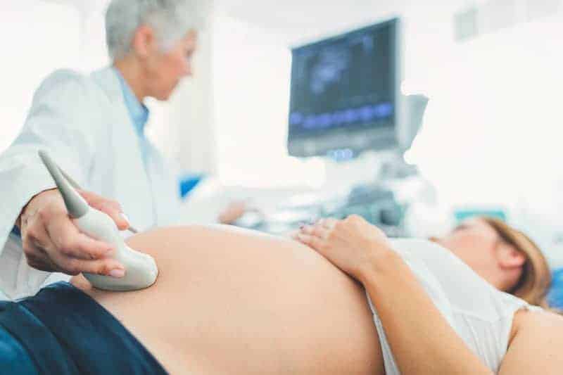 A doctor uses an ultrasound transducer on a pregnant woman's belly in a bright medical office.