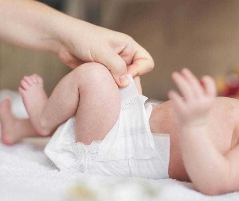 Close-up of a person's hand opening the side tab of a white diaper on a baby lying on their back.