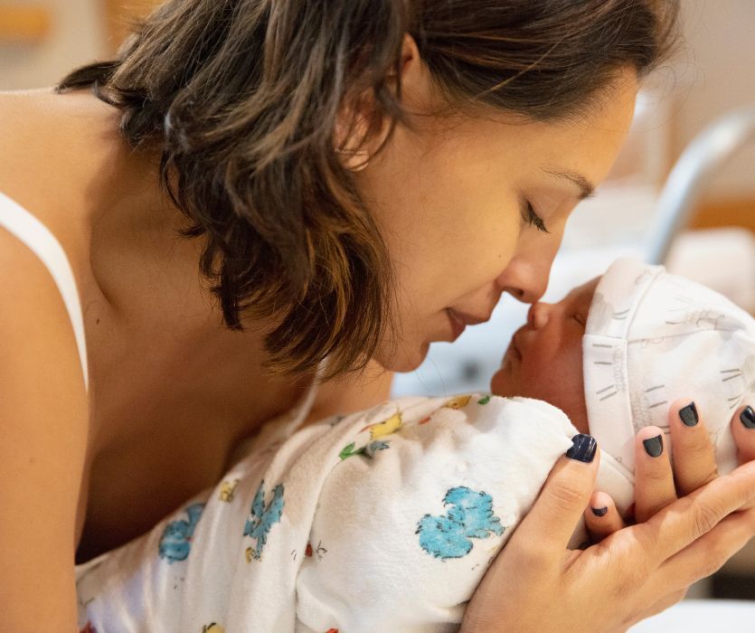 A woman leans down to press her nose against the face of a swaddled newborn baby in a hospital setting.