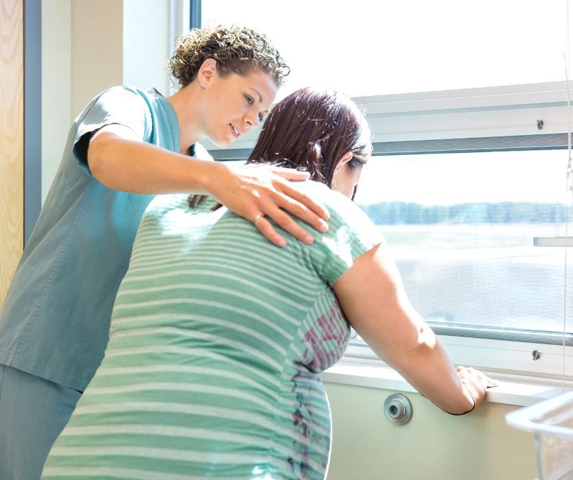 A doula in scrubs places a hand on the back of a pregnant woman leaning against a windowsill during labor.