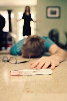 A positive pregnancy test on the floor near a fainted man while a woman stands in the background.
