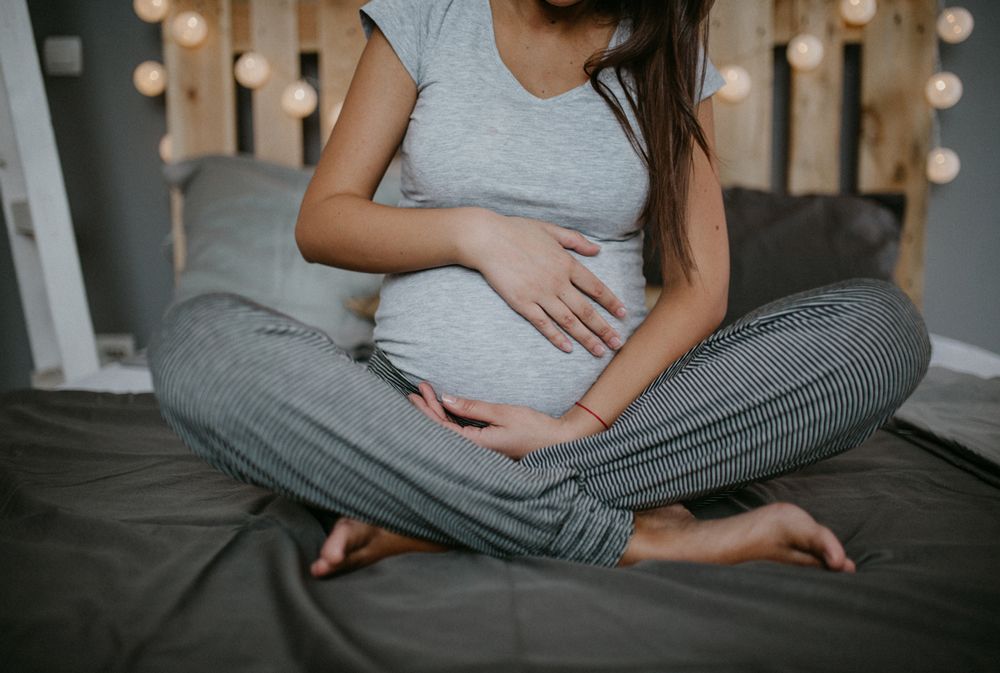 Pregnant Moira sits cross-legged on a bed in a gray top and striped pants, cradling her belly beside soft bedroom lights during a calm moment.
