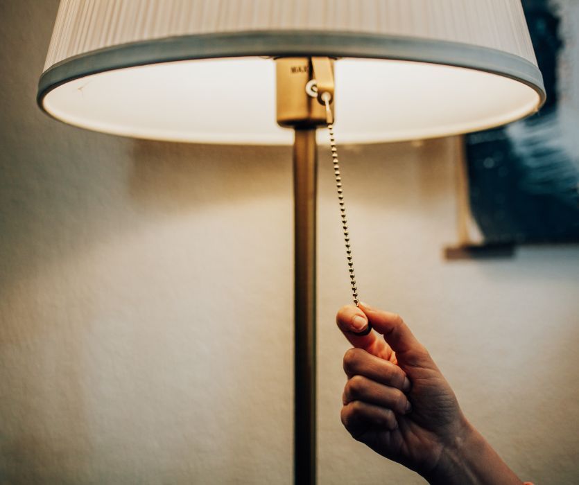 A hand reaching up to pull the beaded metal chain of a glowing floor lamp.