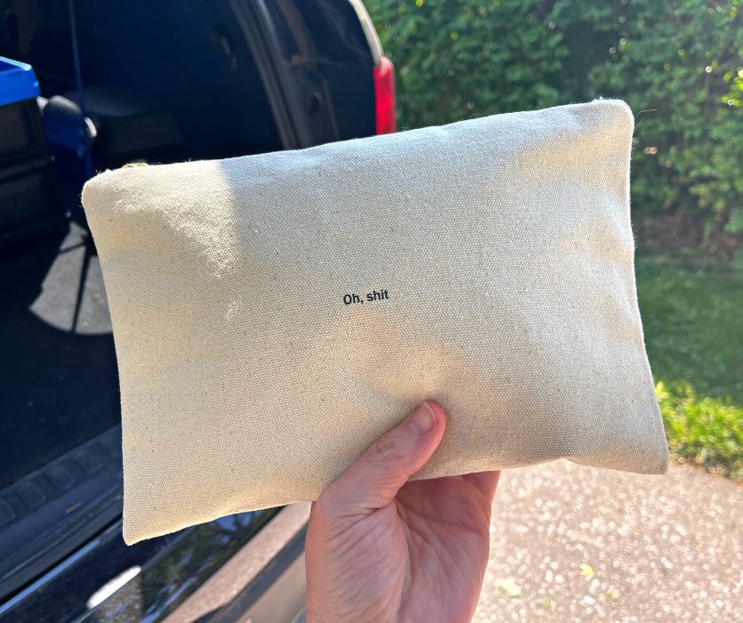 A hand holds a canvas car emergency kit pouch with the text Oh, shit printed on the front.