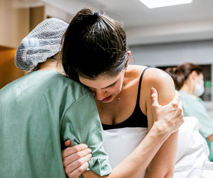 A medical professional in scrubs and a hairnet supports a woman in labor during an epidural procedure.