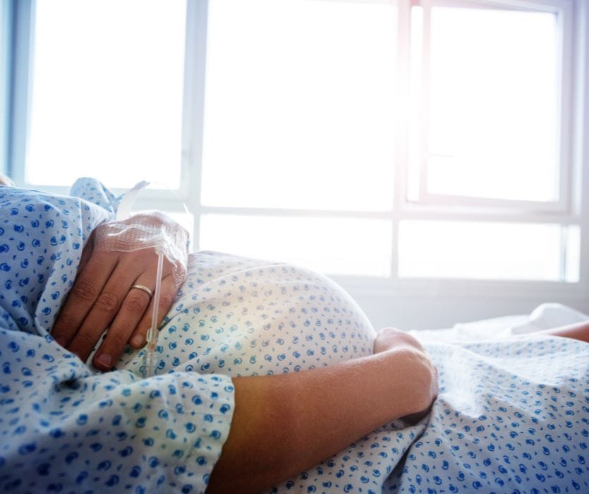 A pregnant person in a hospital gown lies in bed with an IV line in their hand while resting a hand on their stomach.