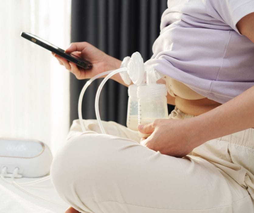 A woman sits cross-legged using a double electric breast pump while holding a smartphone.