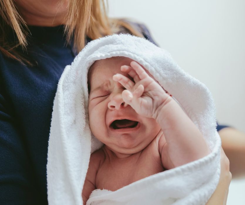 Upset crying newborn wrapped in a white towel being held by a person in a blue shirt.