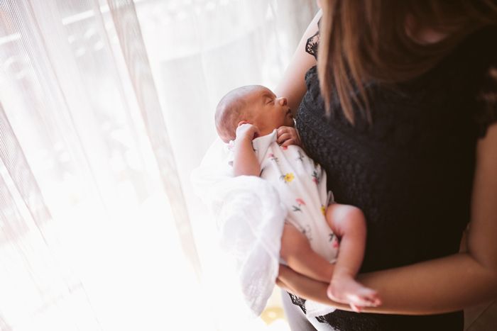 A mother holding a sleeping newborn baby wrapped in a white floral swaddle near a bright window.