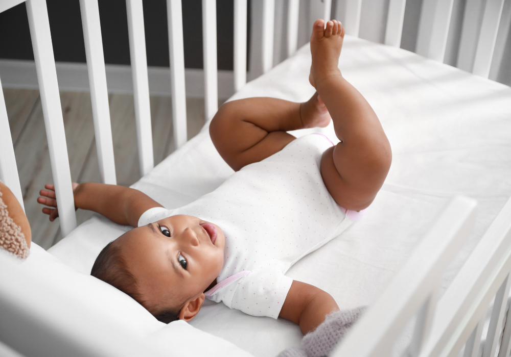 A baby wearing a white onesie lies on their back in a white crib, looking up and smiling.