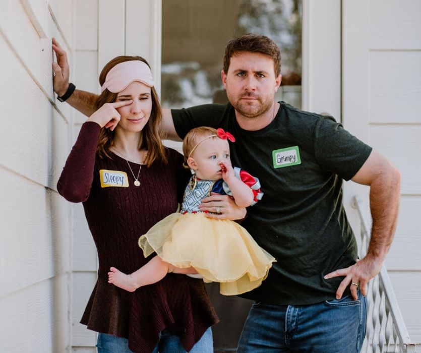 A man and woman in Sleepy and Grumpy tags hold a baby dressed as Snow White for a family costume.