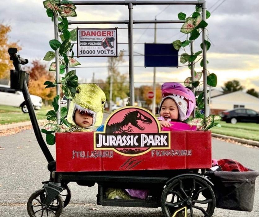 Two children in dinosaur costumes sitting in a wagon decorated like a Jurassic Park enclosure.