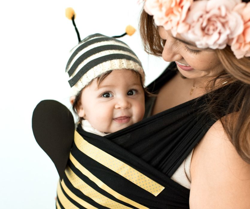 A mother smiles at her baby dressed in a bumblebee costume while being carried in a black and yellow striped baby wrap.