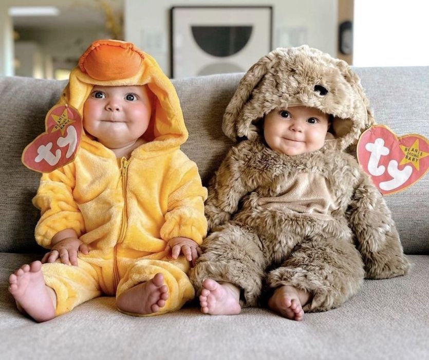 Two babies sitting on a couch wearing plush duck and puppy costumes with giant Beanie Baby heart tags attached.