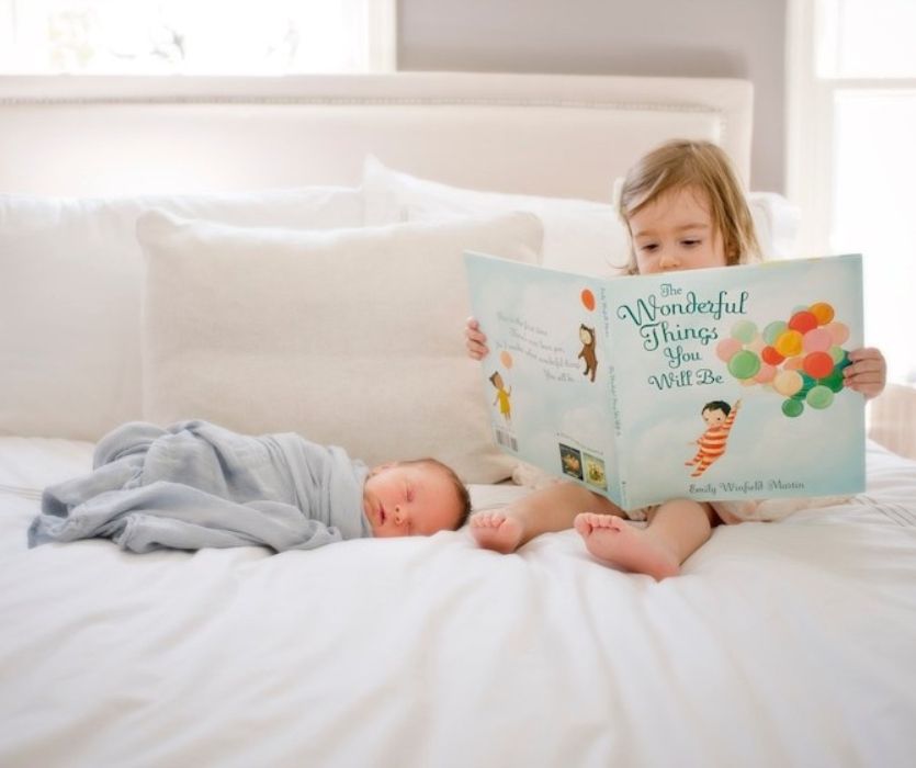 A young girl sits on a bed reading a book to a swaddled, sleeping newborn baby.