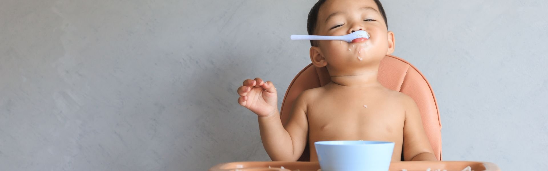A toddler in a high chair eating a meal with a spoon and blue bowl.