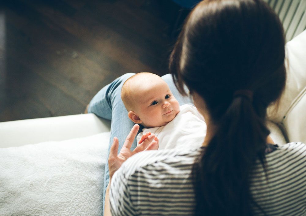 A wide-eyed infant in a white onesie lies on a woman's lap and gazes up at her.