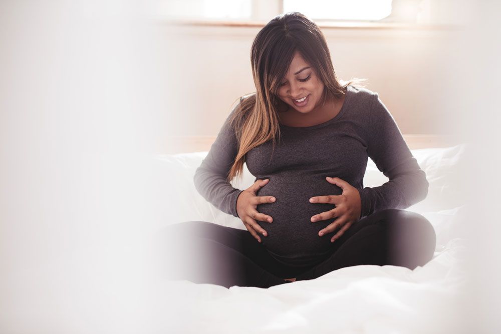A smiling pregnant woman sitting on a bed and holding her baby bump.