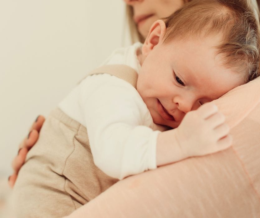 A 14-week-old infant in tan overalls resting their head on a person's shoulder.