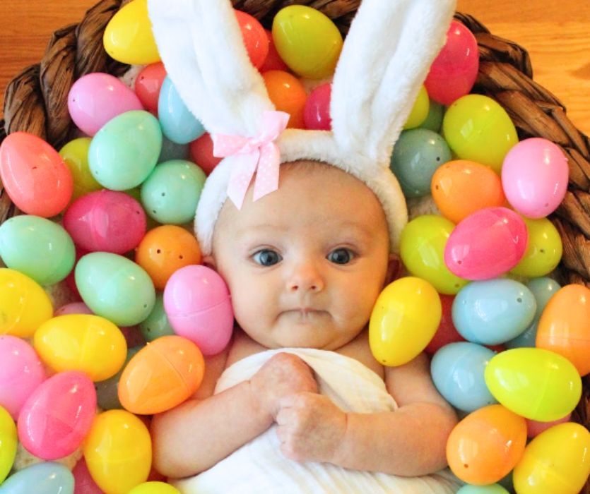 A baby wearing white bunny ears with a pink bow lies in a basket surrounded by colorful plastic Easter eggs.
