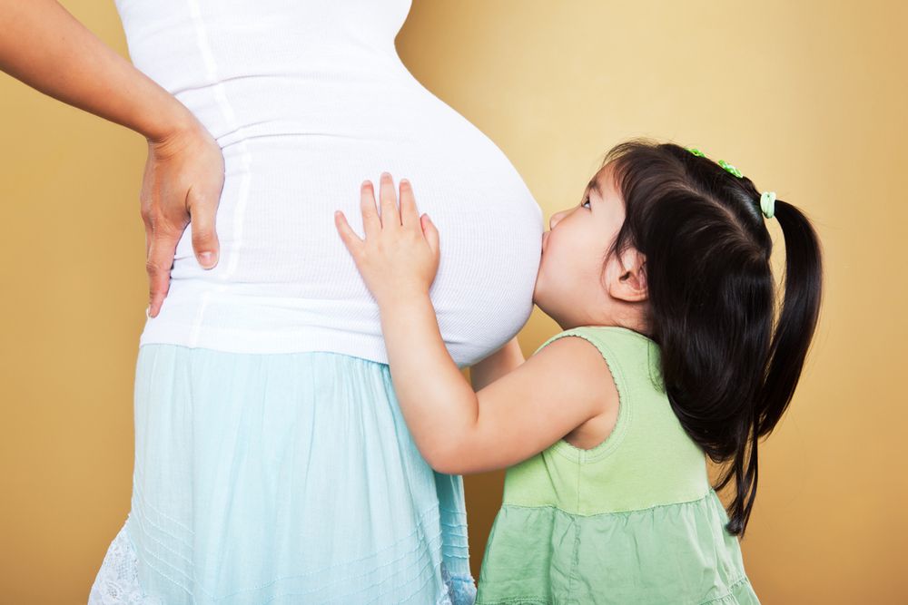 Young girl kissing a woman's pregnant belly against a gold background.
