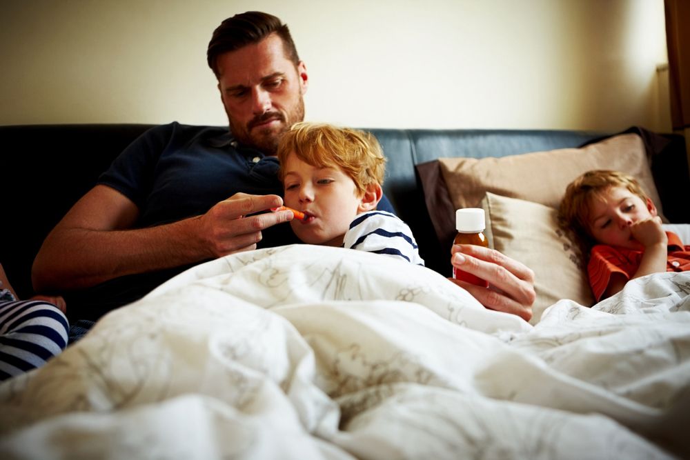 A father gives liquid medicine to his sick son in bed using an oral syringe while another child rests nearby.