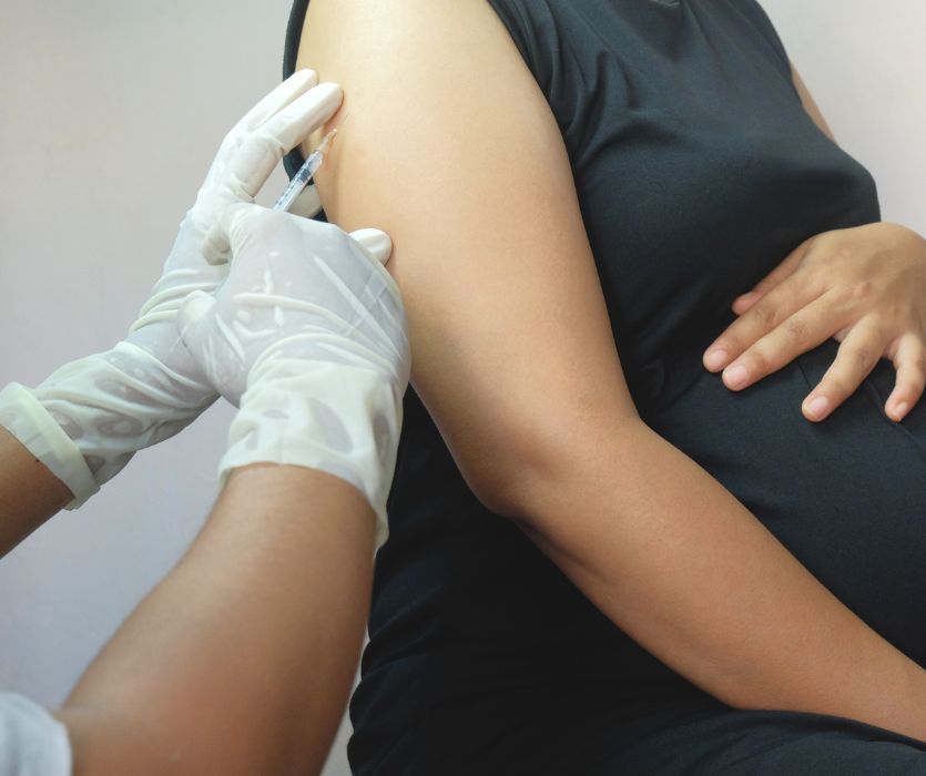 A pregnant woman in black receives a flu vaccine injection in the upper arm from a clinician wearing white gloves during pregnancy.