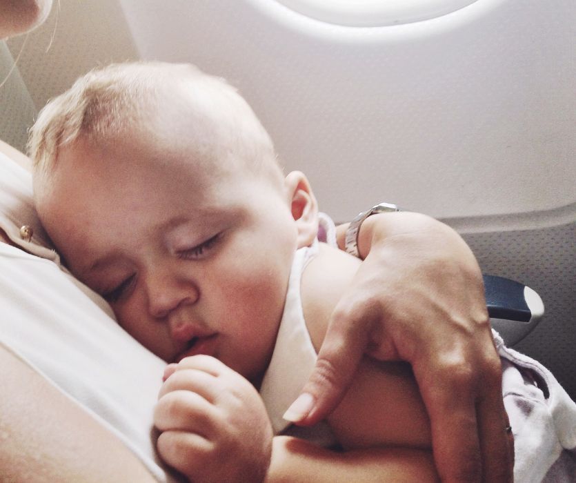 A close-up of a sleeping baby being held in an adult's arms on an airplane next to a window.