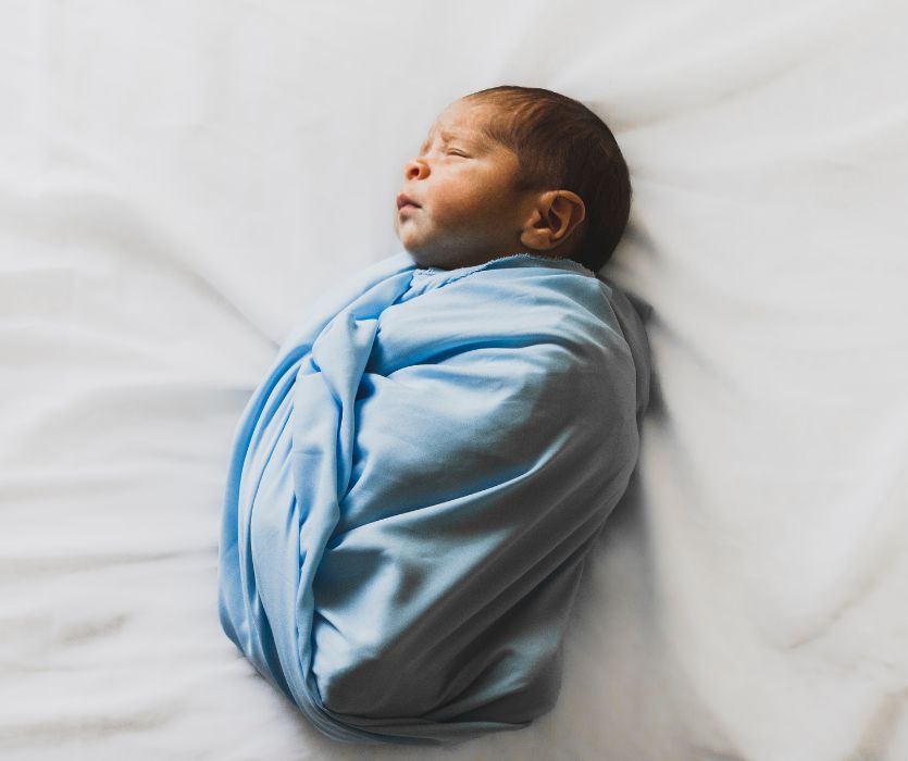 A sleeping newborn baby wrapped in a light blue swaddle blanket, lying on a white sheet.