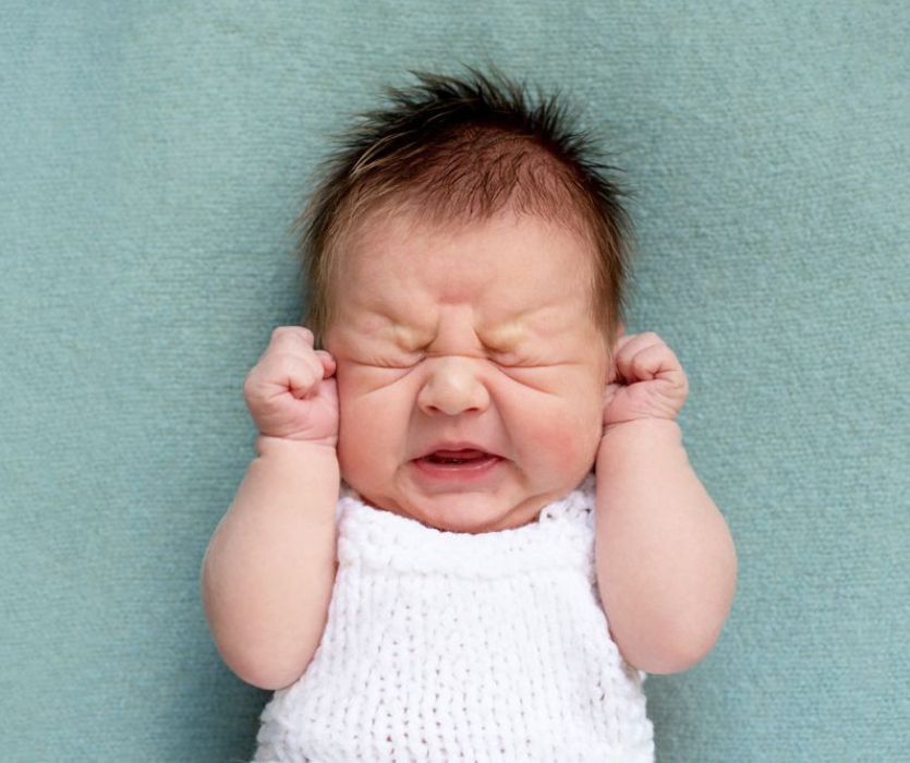 Close-up of a crying newborn baby with eyes squeezed shut and fists clenched near their ears.