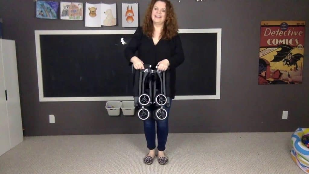 A woman stands in a room holding a GB Pockit stroller folded into its most compact, square configuration.
