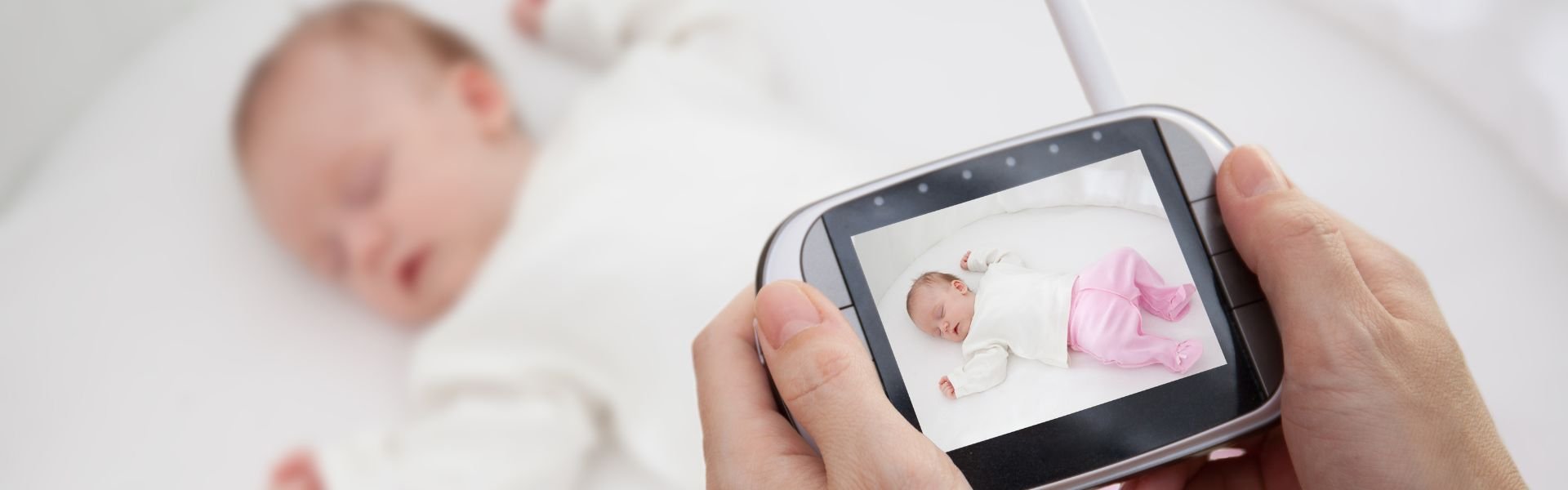 A person's hands holding a video baby monitor showing a sleeping infant. A blurred baby sleeps in the background.