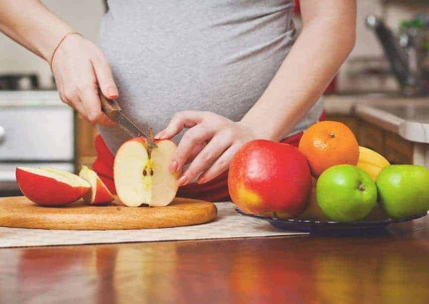 A pregnant person slicing a red apple on a wooden board next to a bowl of fresh fruit.