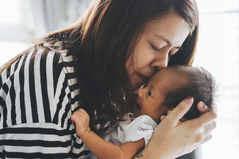 A woman in a striped shirt kisses the forehead of a newborn baby held in her arms.
