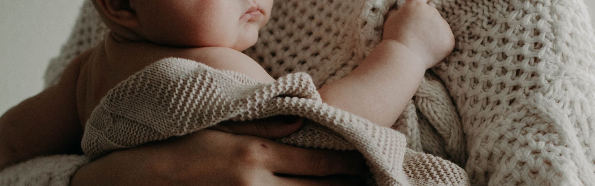 Close-up of a baby held in an adult's arms, draped in a soft, cream-colored knit blanket.